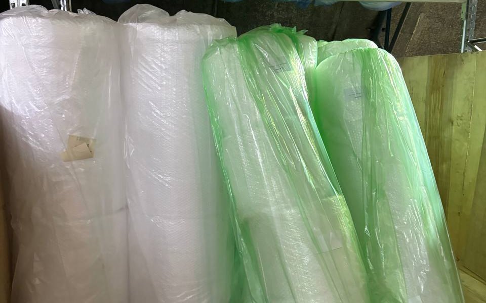 Large rolls of clear and green-tinted bubble wrap packing material standing on a wooden floor under a shelf