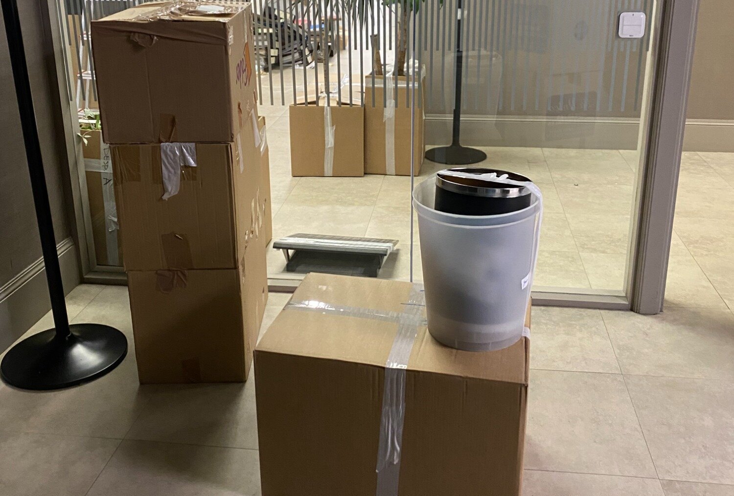 Taped cardboard moving boxes and a bin stacked on a tiled floor next to a glass partition, ready to be carried out