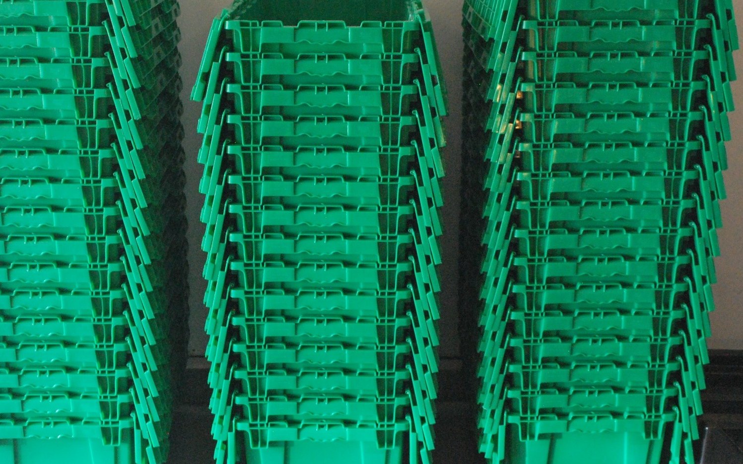 Three tall stacks of green reusable plastic moving crates with attached lids lined up against a garage wall